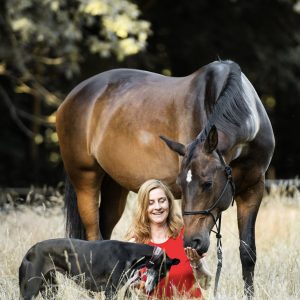 Frau sitzt auf dem Boden, Pferd und Hund stehen rechts und links neben ihr und berühren mit ihrern Nüstern bzw. Schnauze die offenen nach oben gedrehten Hände